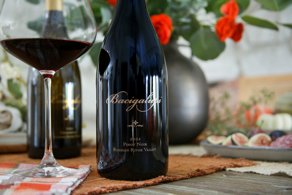 Photo of a 2022 Bacigalupi Pinot Noir on a table with flowers behind and a partially filled glass to the side.